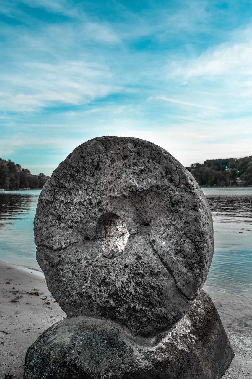 Weathered circular millstone beside lake water and a wooded shoreline.