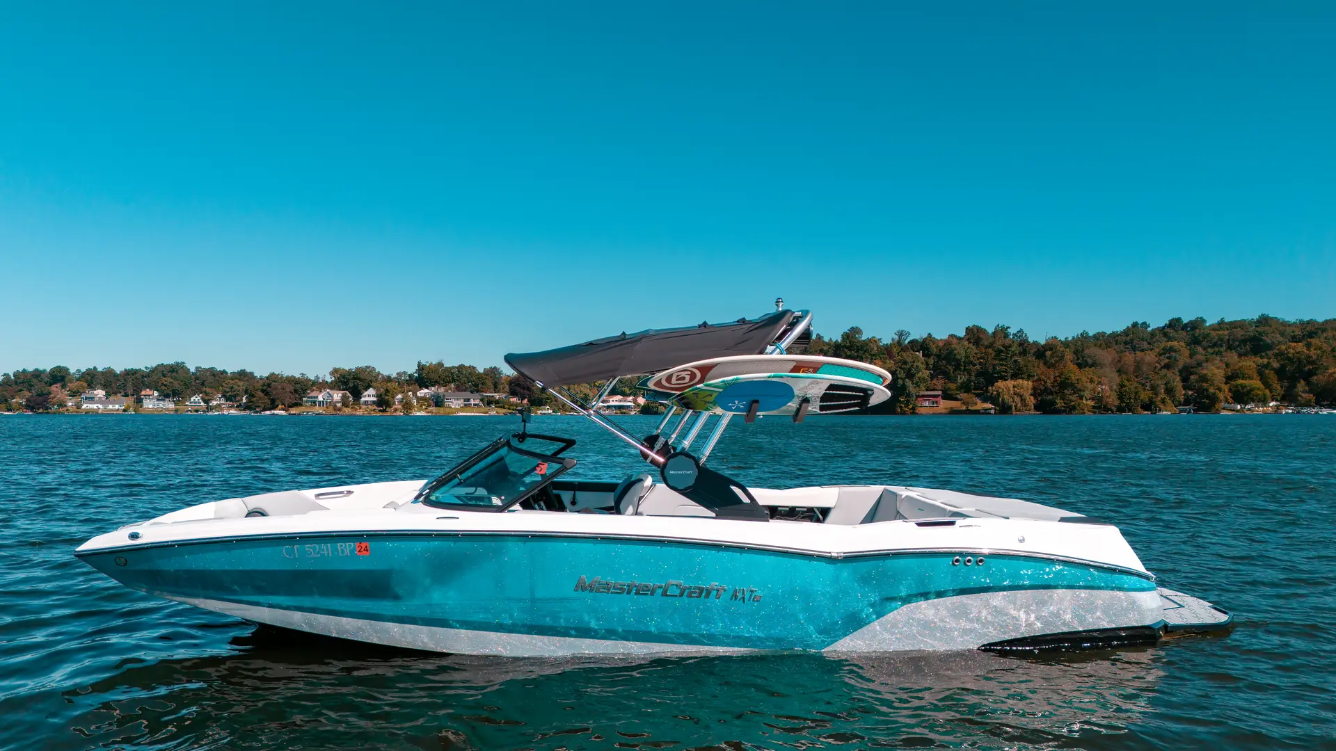 A wake boat on calm water on Candlewood Lake, Connecticut, with the wooded shoreline behind it.